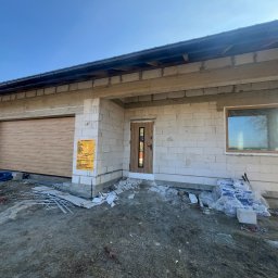 Okna LaVico Piotr Wontorczyk - Nowy garaż z drewnopodobną bramą segmentową, drzwi wejściowe z przeszkleniem i okno w surowym stanie. Widoczne materiały budowlane i tablica informacyjna.