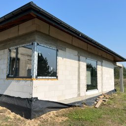 Okna LaVico Piotr Wontorczyk - Surowy stan budynku z zamontowanymi oknami PCV w czarnych ramach. Widoczne ocieplenie wokół okien i folia fundamentowa. Tło stanowi błękitne niebo i zieleń.