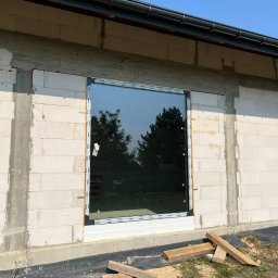 Okna LaVico Piotr Wontorczyk - Montaż okna PCV w surowym budynku z betonu komórkowego. Widoczne drewniane podpory i folia fundamentowa. Okno z folią ochronną na ramie.