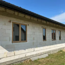 Surowy budynek z jasnego bloczka z zamontowanymi oknami o drewnianej ramie. Widoczne świeże ślady zaprawy. Czarny dach i błękitne niebo w tle.