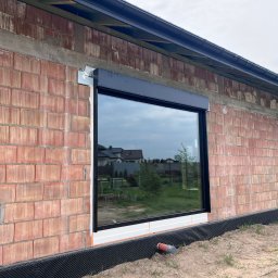 Okna LaVico Piotr Wontorczyk - Czarne okno PCV z roletą zewnętrzną w surowej ścianie z cegły. Widoczne elementy montażowe i fragment instalacji. W tle domy i zieleń.