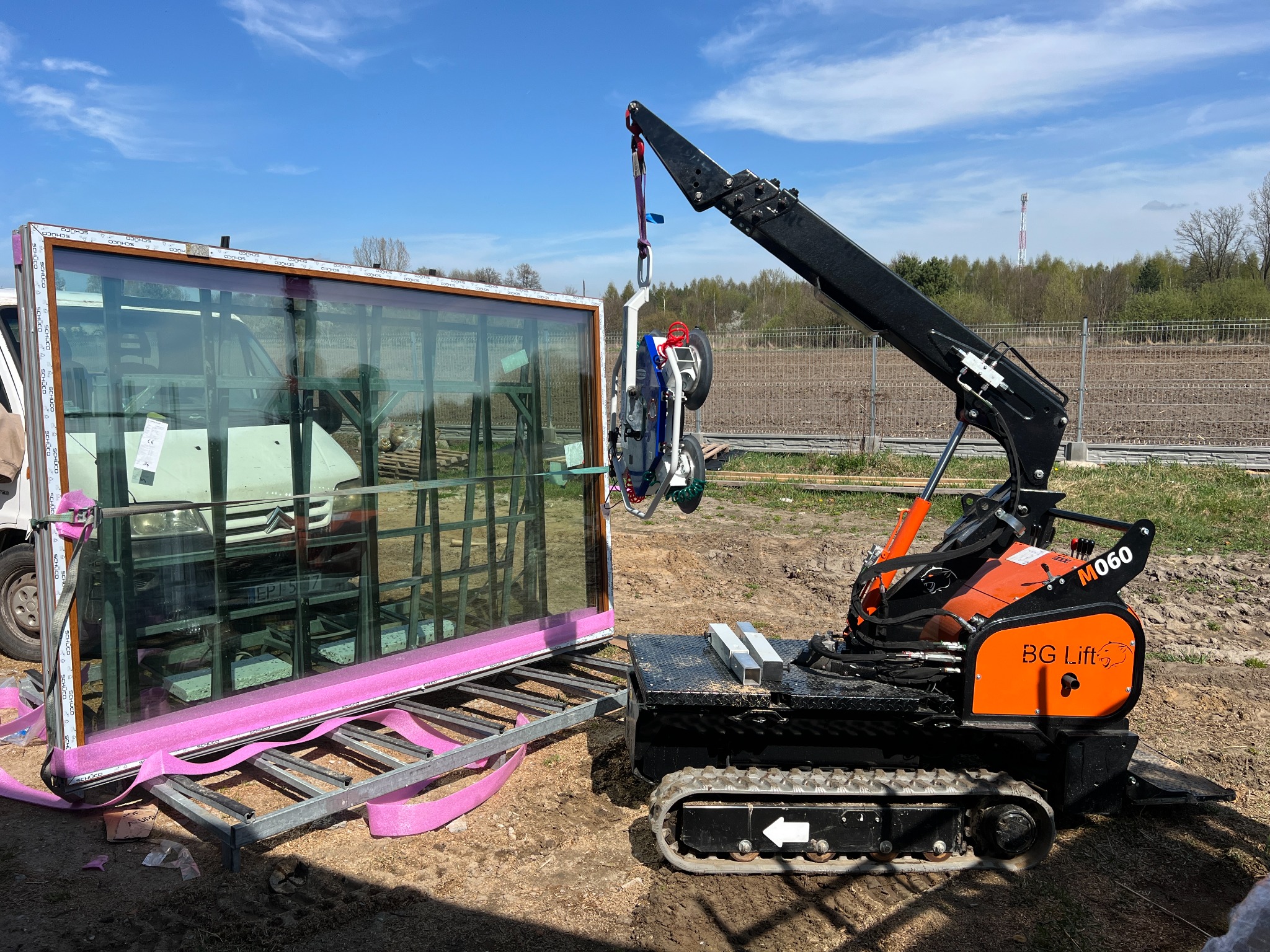 Montaż okna PCV za pomocą mini żurawia BG Lift M060 na gąsienicach. Okno zabezpieczone różową pianką, w tle widoczny samochód dostawczy i częściowo ogrodzenie.