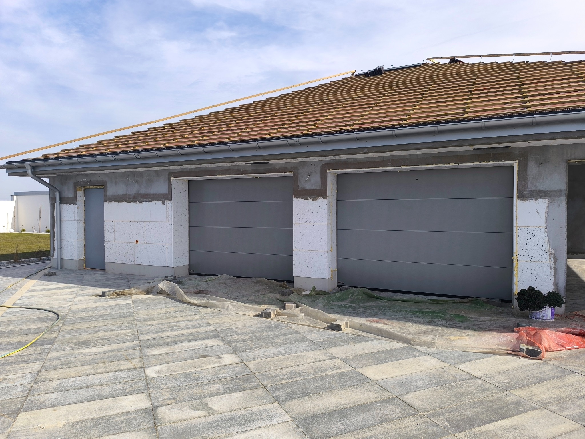 Garaż dwustanowiskowy w trakcie budowy: szare bramy segmentowe, surowy stan ścian, ułożona kostka brukowa. Widoczny fragment dachu z konstrukcją pod dachówkę.