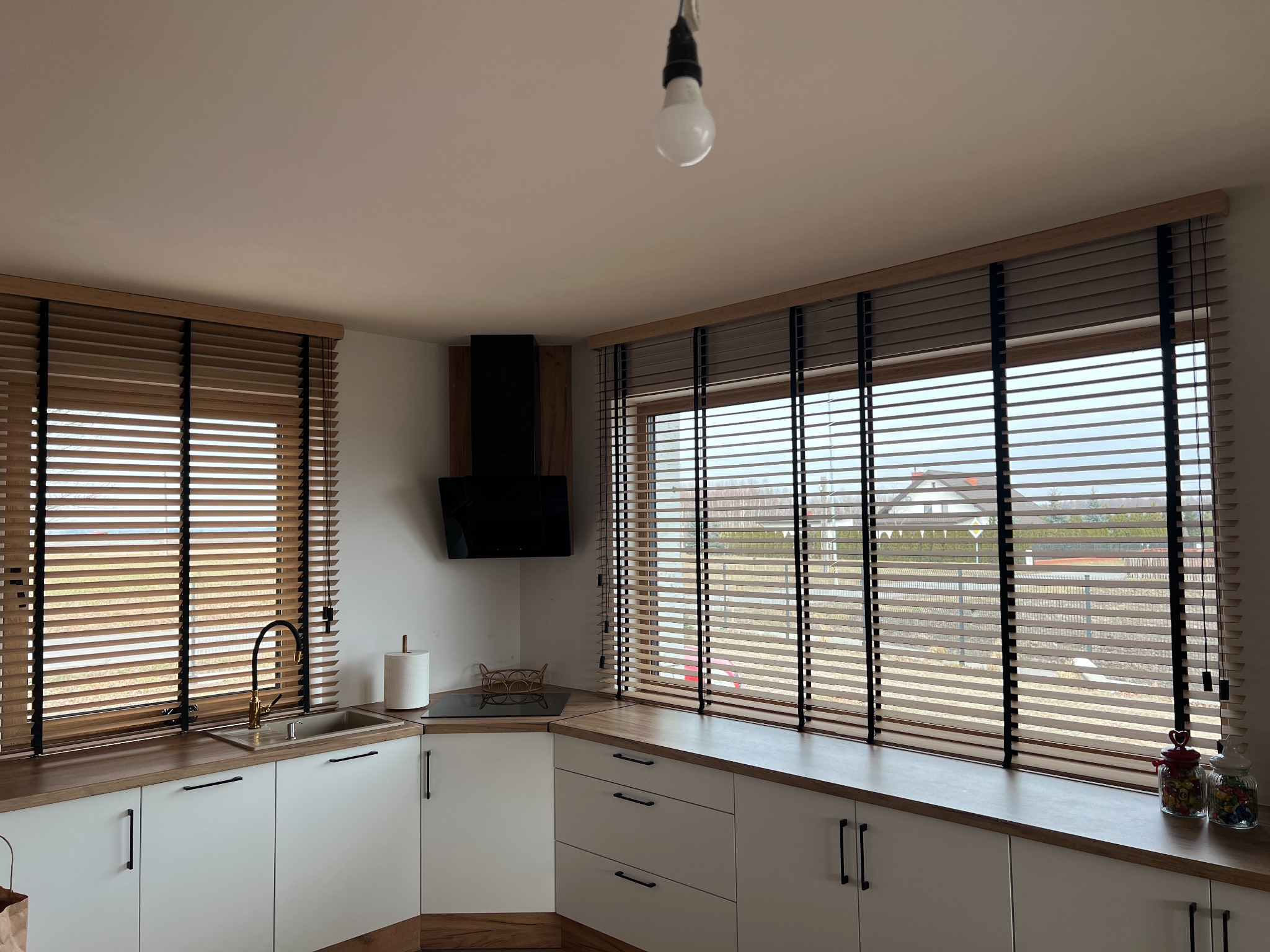 Kuchnia z drewnianymi żaluzjami w oknach, jasne szafki i drewniany blat. Widok na zewnątrz przez okno. Minimalistyczny design.