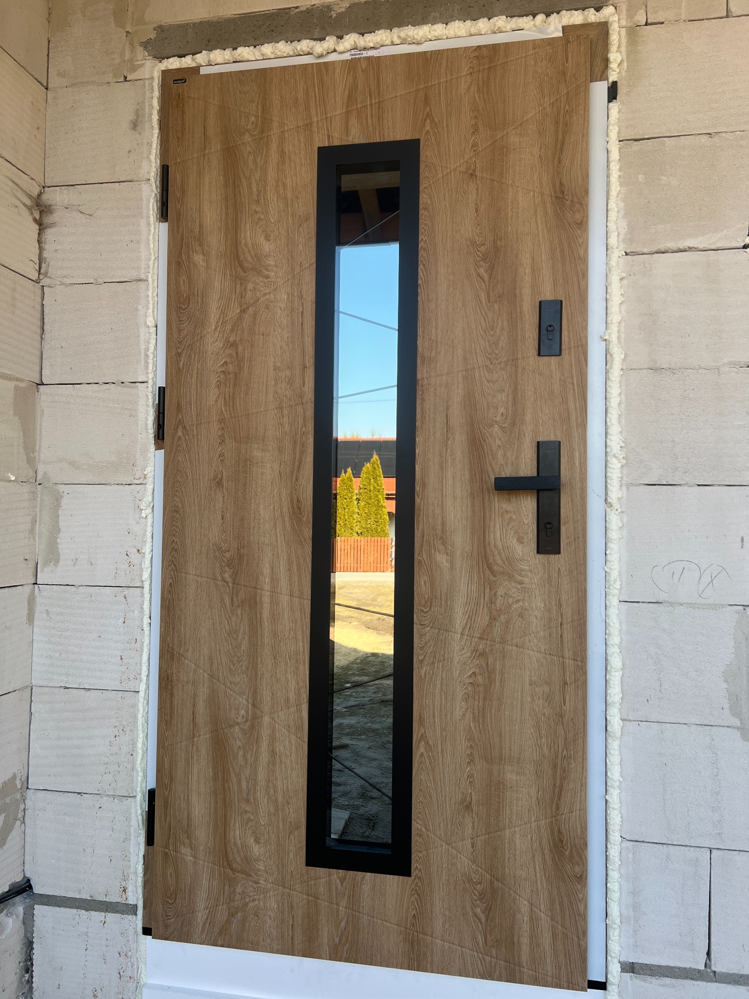 Zamontowane drzwi zewnętrzne z drewnopodobnym panelem i długim, wąskim przeszkleniem w czarnej ramie, osadzone w surowej ścianie z betonu komórkowego.