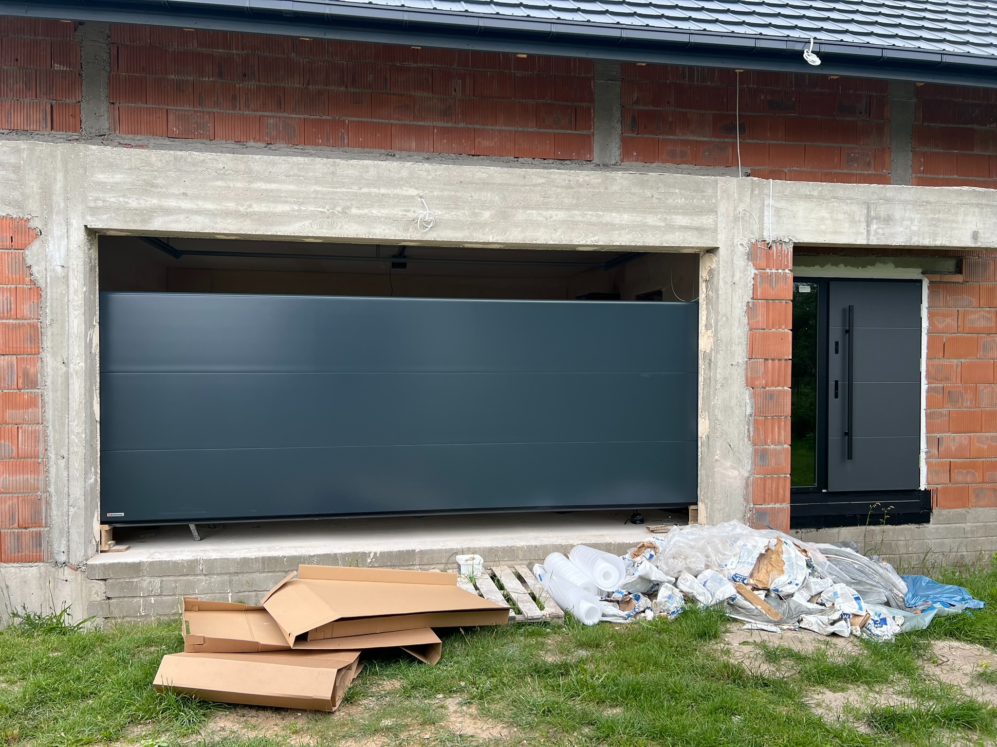 Ciemnoszara brama garażowa segmentowa w surowym otworze budynku z cegły. Na dole kartony i folie montażowe. Obok drzwi wejściowe w kolorze bramy.