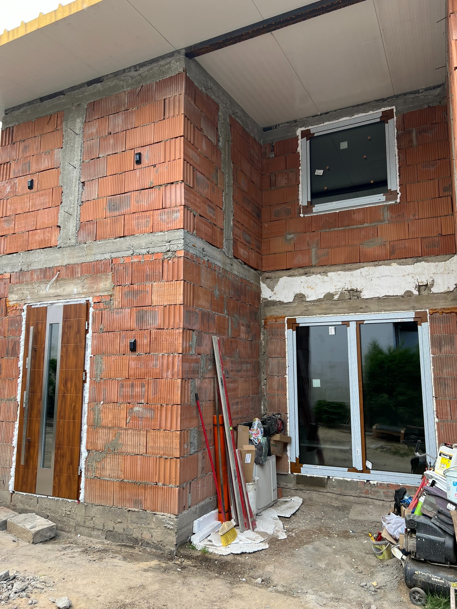 Montaż stolarki okiennej i drzwiowej w surowym budynku z czerwonej cegły. Widoczne okna PCV, drzwi zewnętrzne i narzędzia.