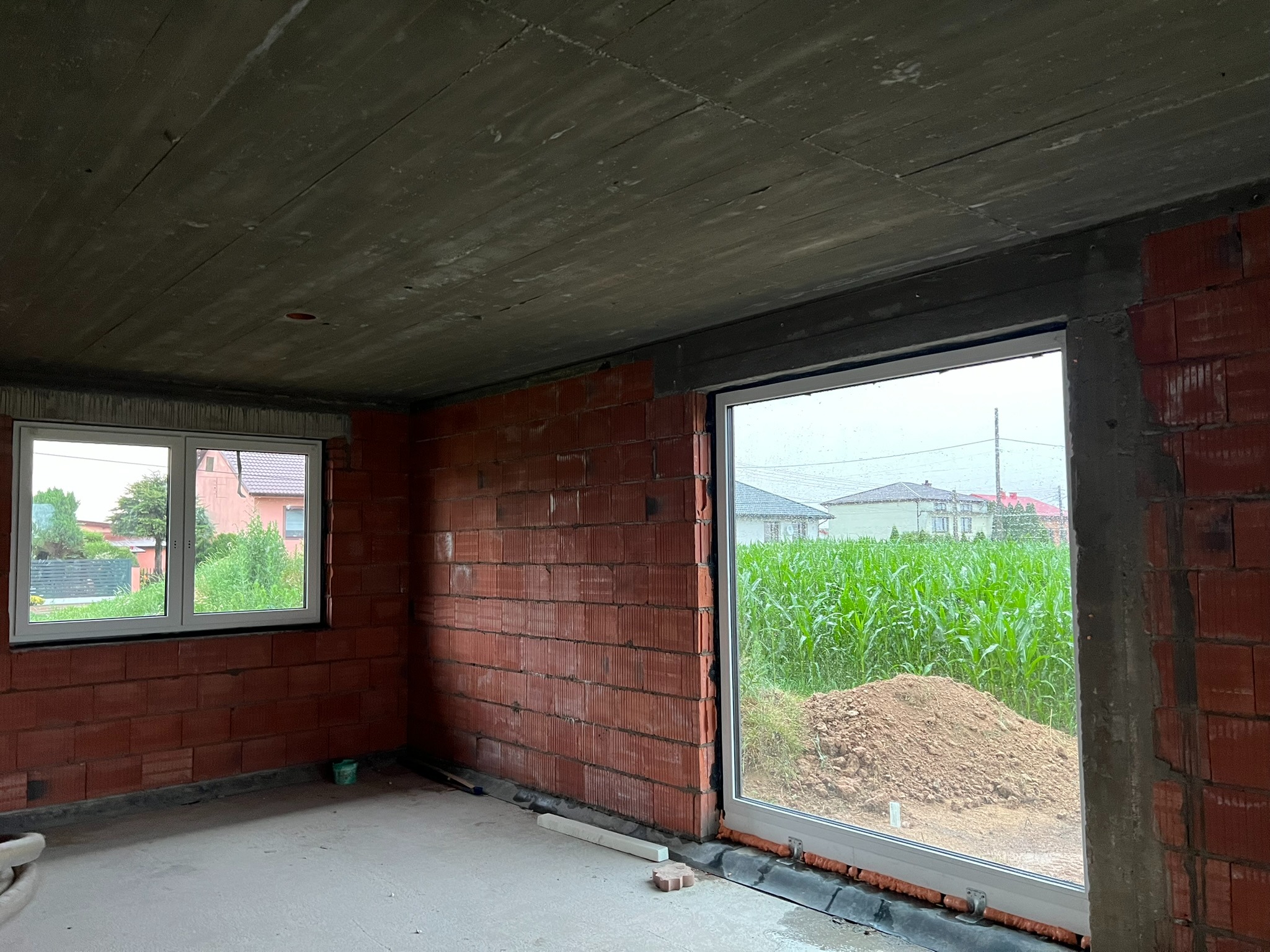 Wnętrze surowego budynku z dwoma oknami PCV; jedno mniejsze, drugie duże z widokiem na zieleń. Ściany z czerwonej cegły, betonowy strop. Na zewnątrz widoczny krajobraz.