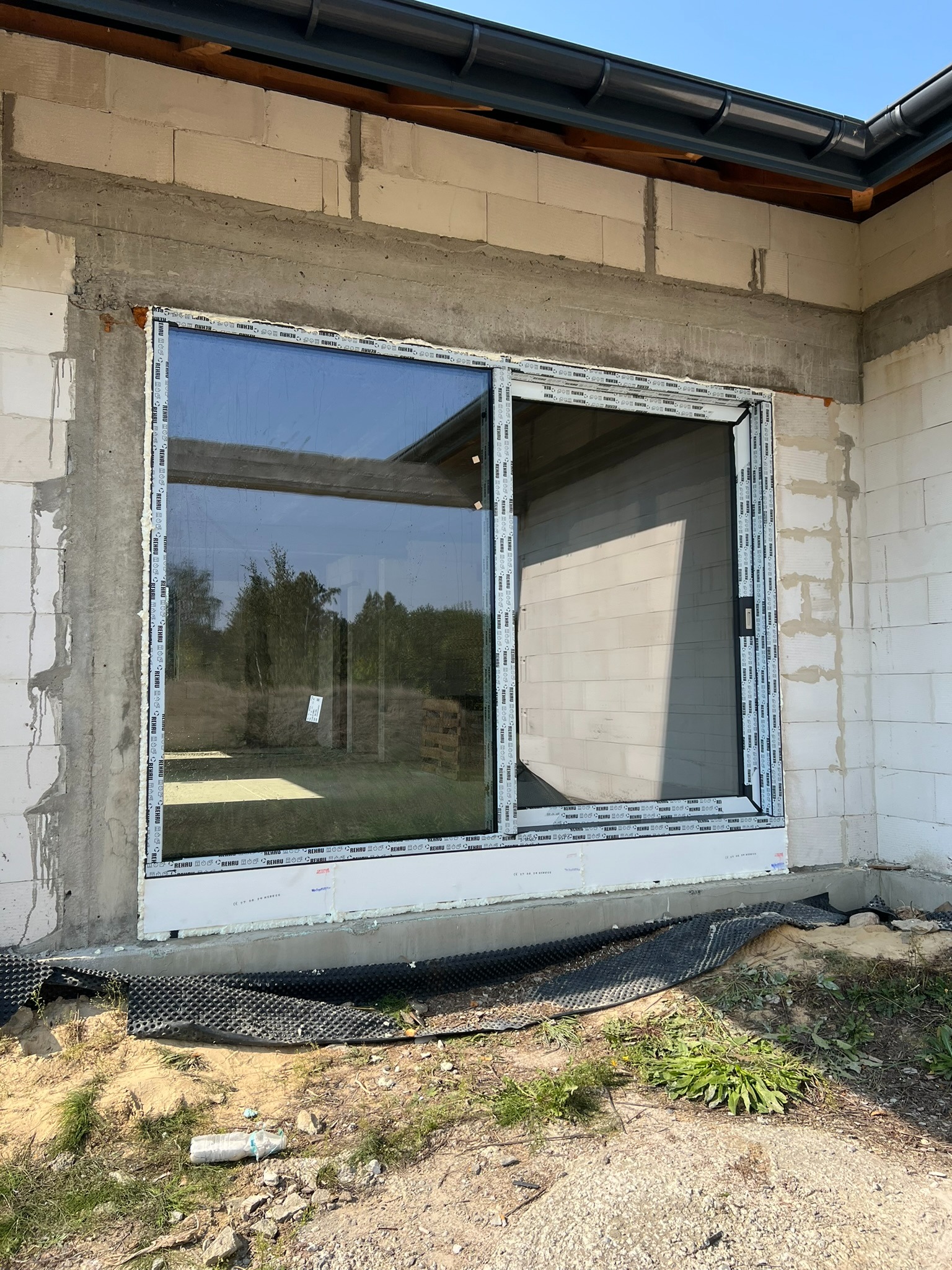 Montaż okna przesuwnego w surowym murze, widoczne detale ramy z logo producenta. Częściowo otwarte skrzydło okienne. Na dole widoczna folia fundamentowa.