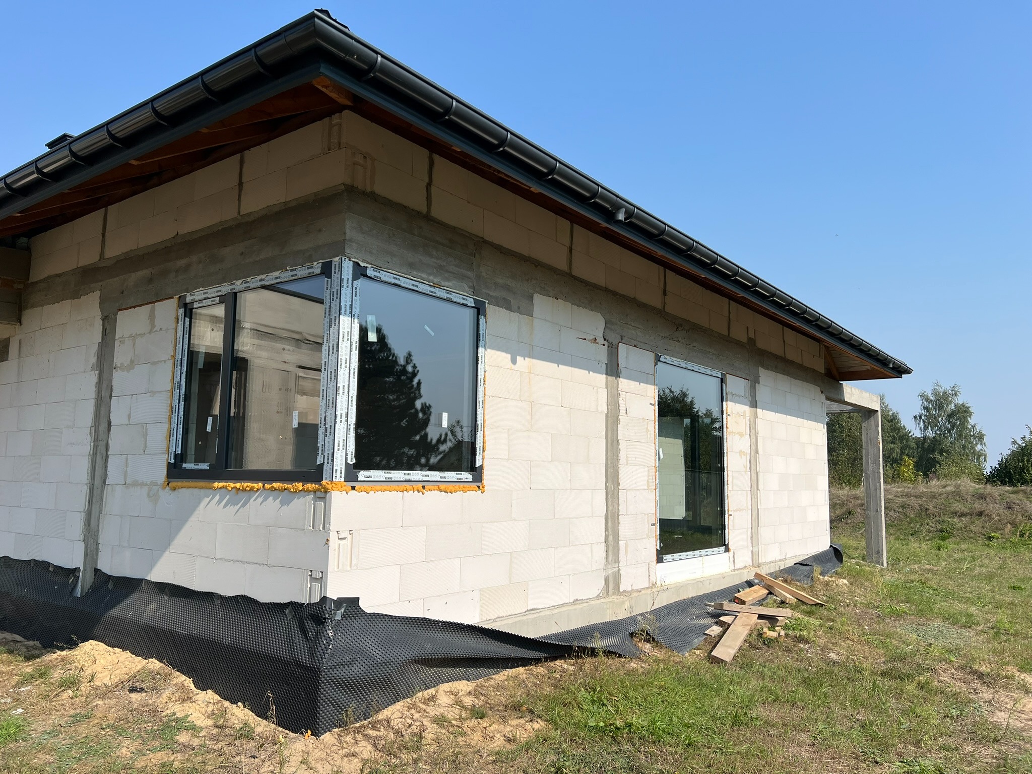 Surowy stan budynku z zamontowanymi oknami PCV w czarnych ramach. Widoczne ocieplenie wokół okien i folia fundamentowa. Tło stanowi błękitne niebo i zieleń.