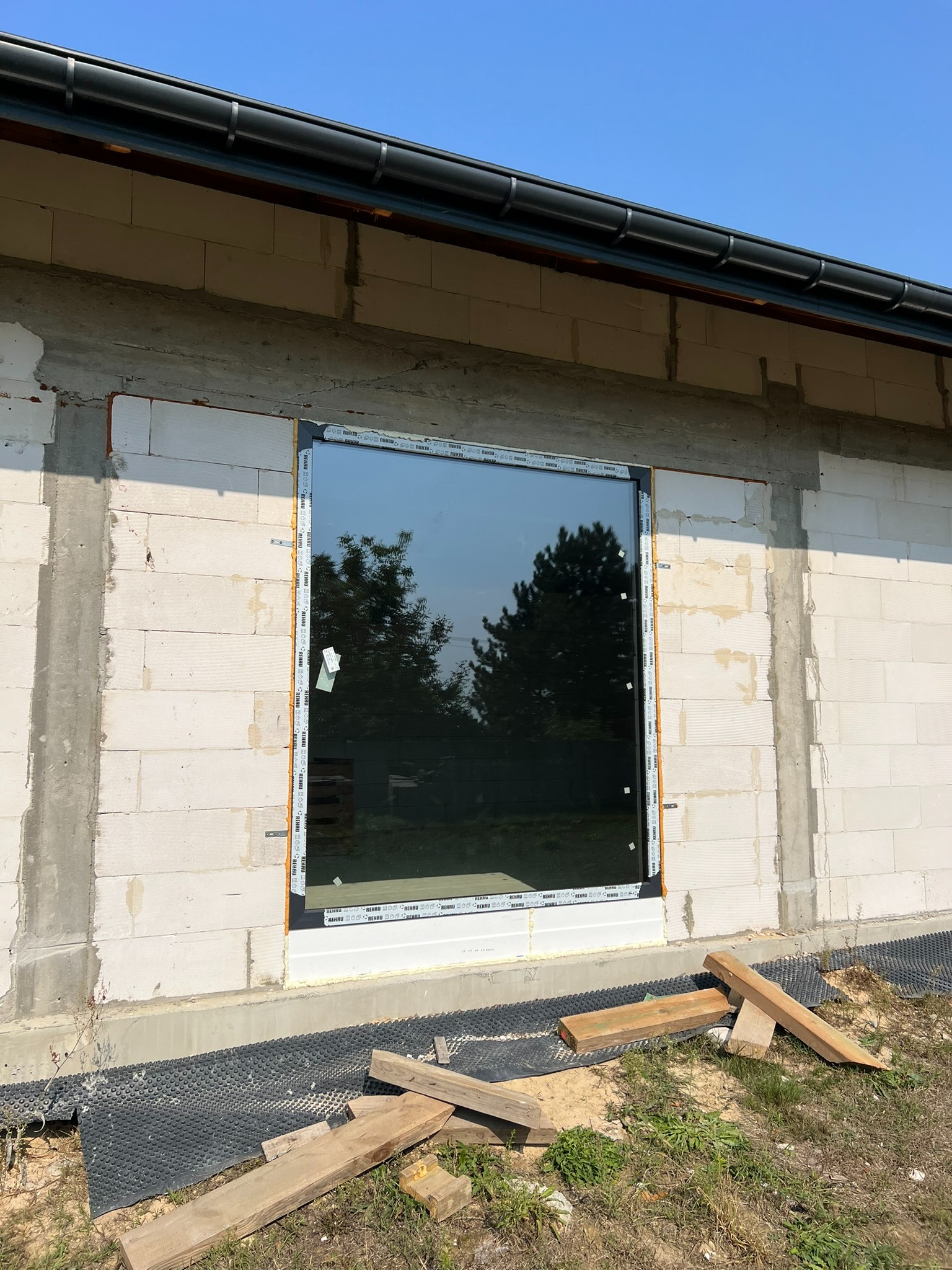 Montaż okna PCV w surowym budynku z betonu komórkowego. Widoczne drewniane podpory i folia fundamentowa. Okno z folią ochronną na ramie.