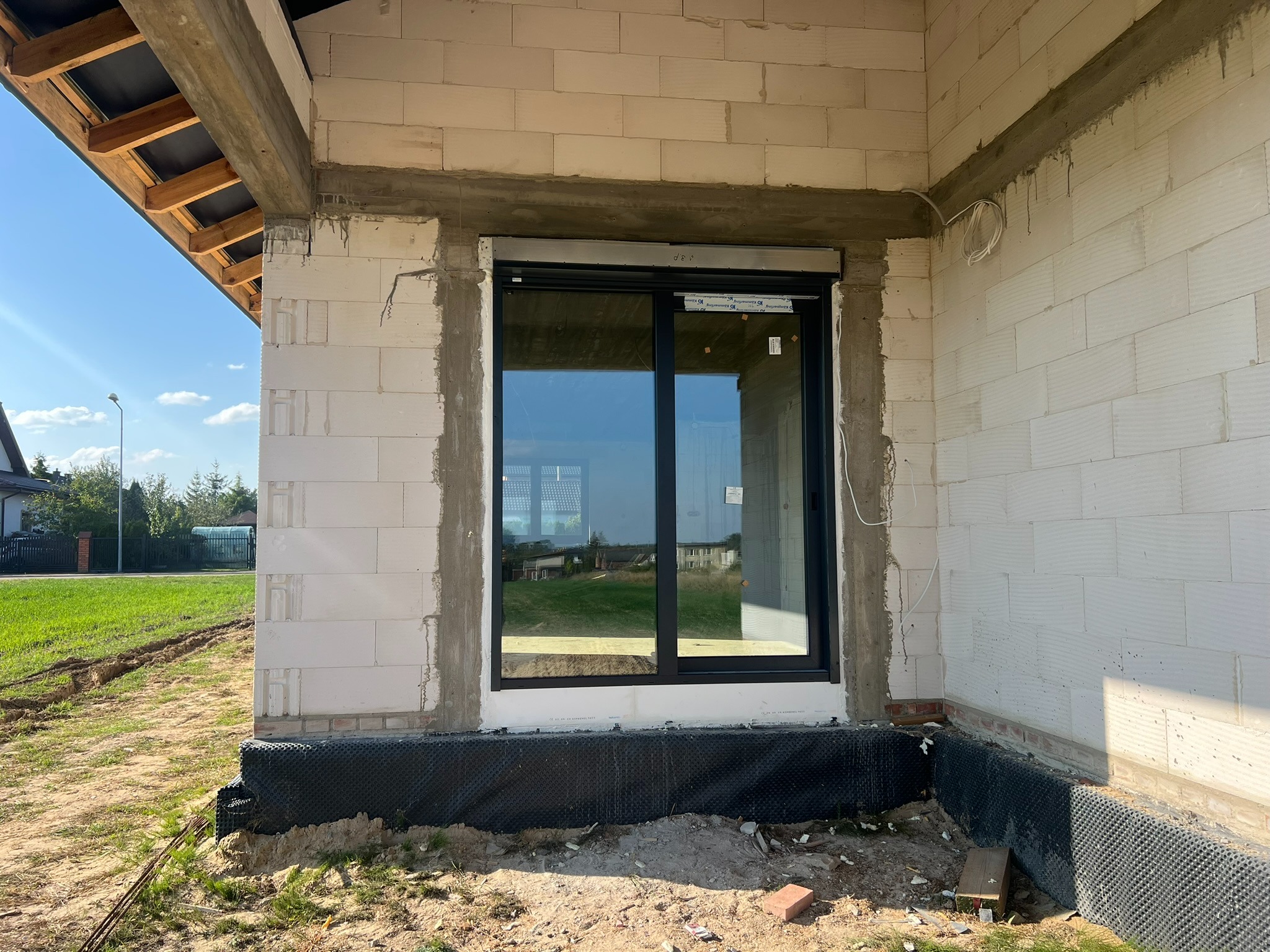 Czarne okno PCV z roletą zewnętrzną w surowym stanie budynku, widok z zewnątrz. Montaż okna z roletą w trakcie budowy domu.