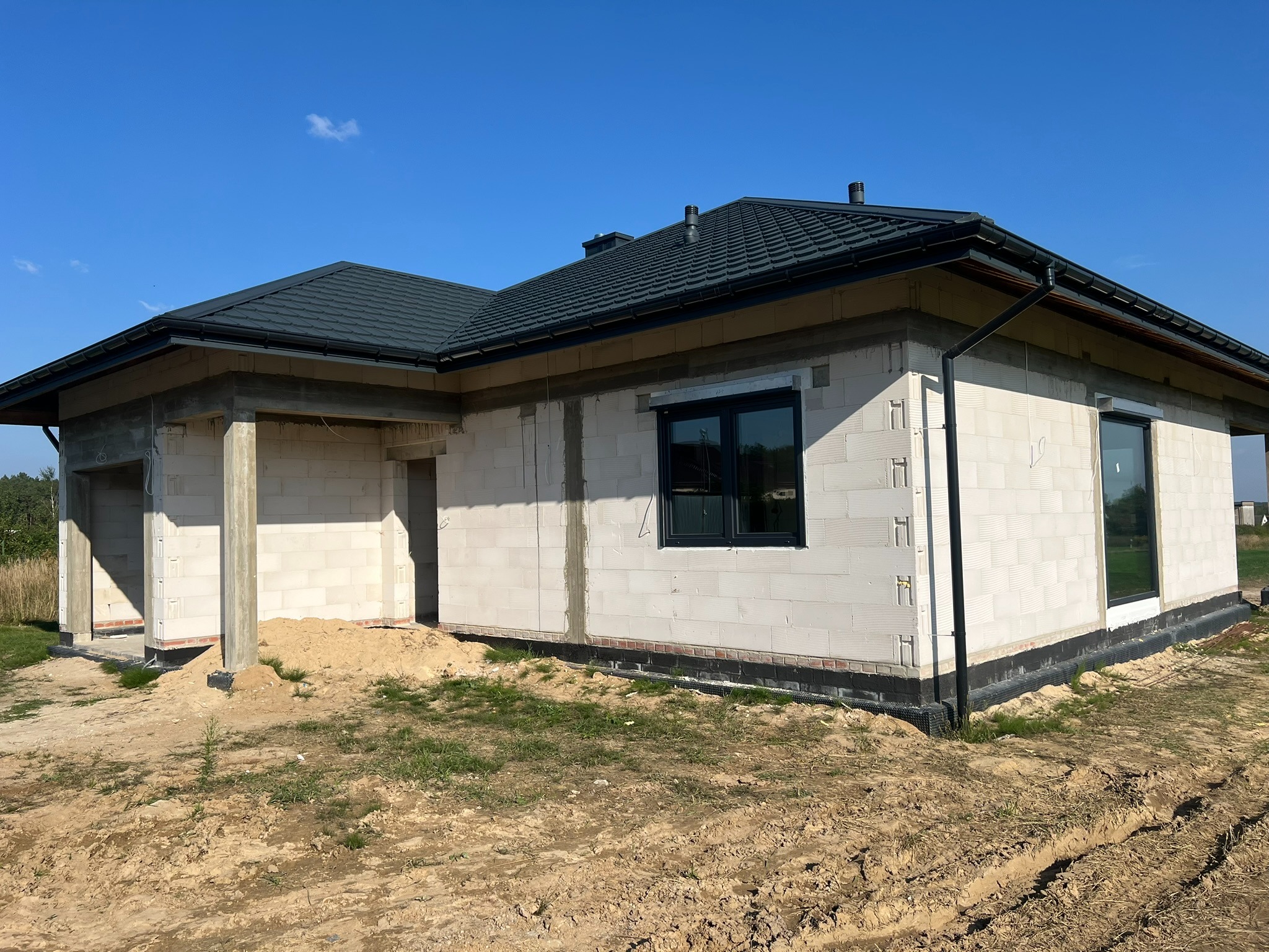 Budynek w stanie surowym otwartym z zamontowanymi oknami PCV w czarnych ramach, widok z zewnątrz, na tle błękitnego nieba.