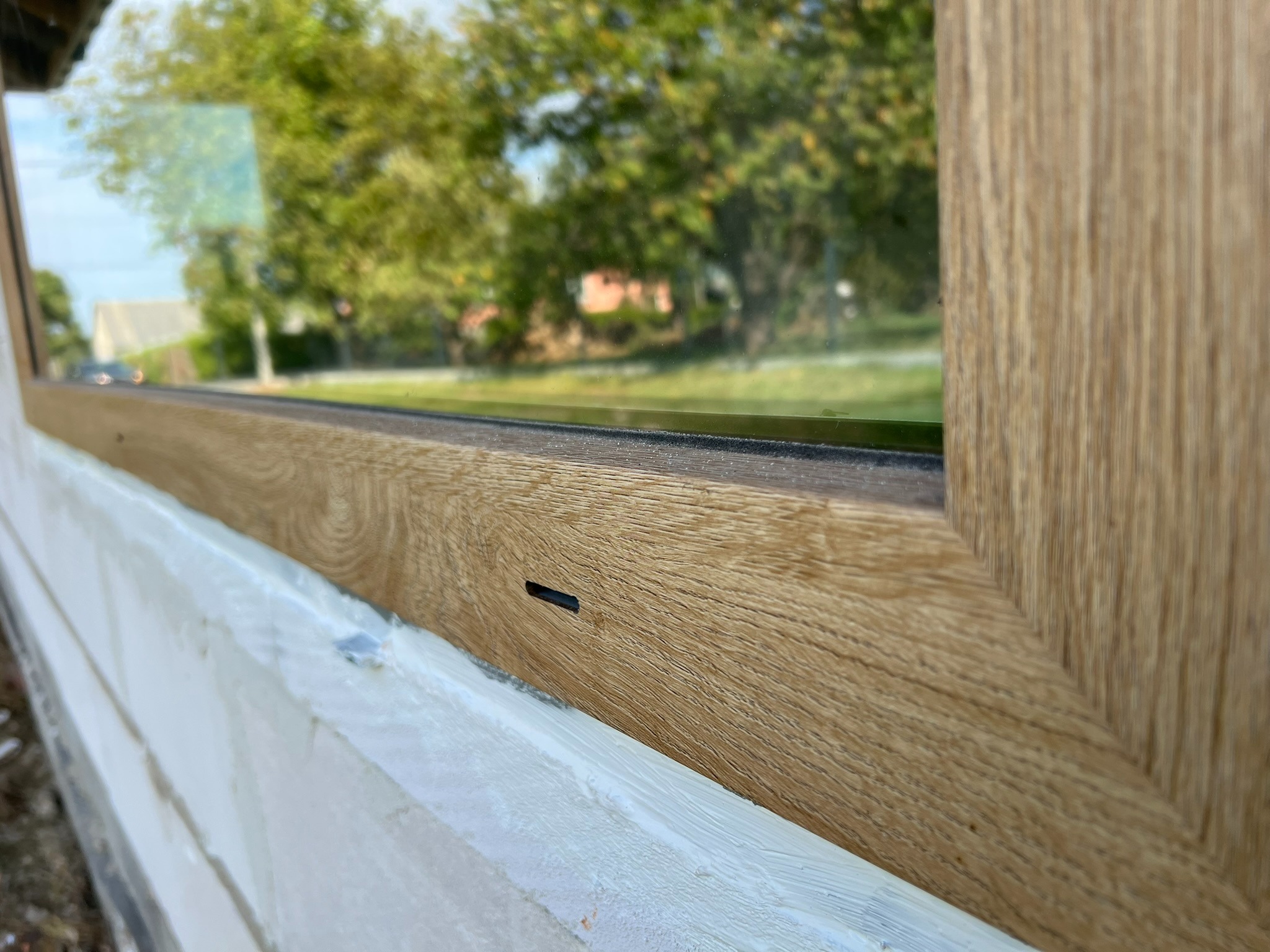 Fragment okna z drewnopodobną ramą, uszczelnionego białą pianką montażową, z widokiem na zieleń za szybą. Ujęcie z bliska, ukazujące detale wykończenia.