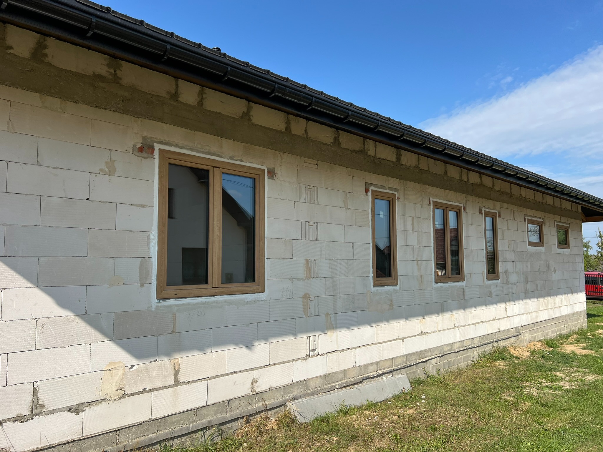 Surowy budynek z jasnego bloczka z zamontowanymi oknami o drewnianej ramie. Widoczne świeże ślady zaprawy. Czarny dach i błękitne niebo w tle.