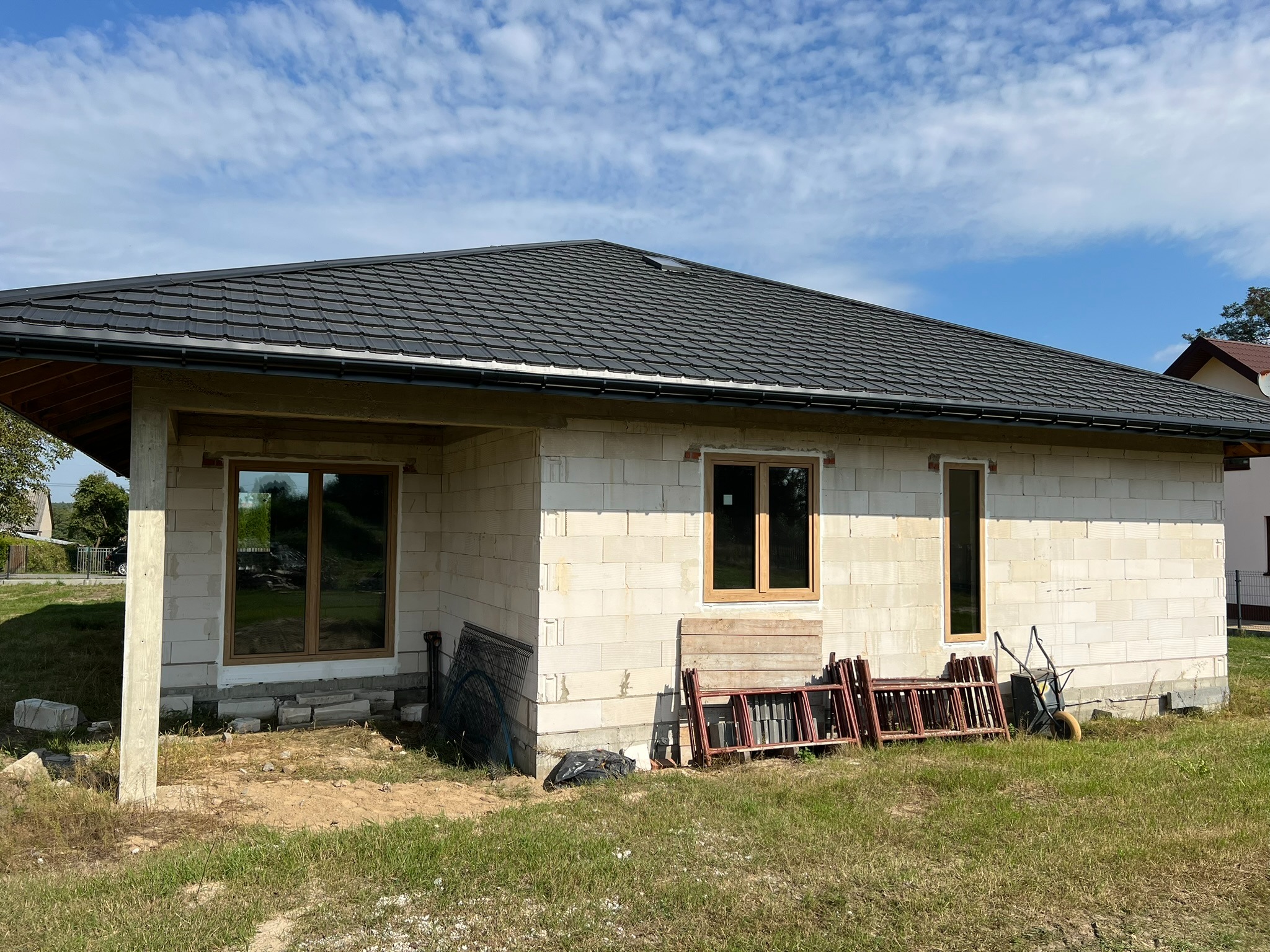Surowy stan domu z oknami w drewnianych ramach, czarnym dachem i narzędziami budowlanymi na trawie. Ściany z białego pustaka, błękitne niebo w tle.