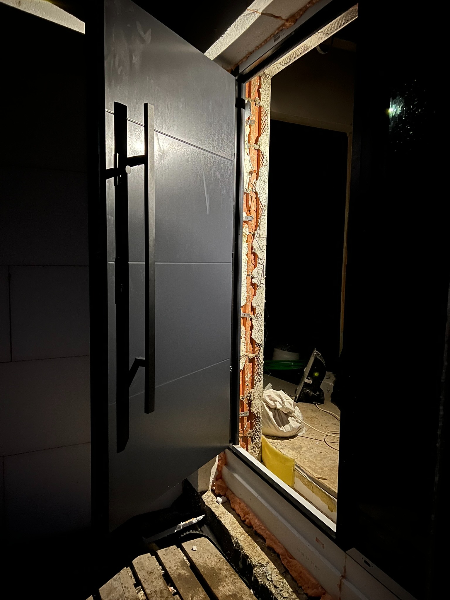 Otwarta, nowoczesna, grafitowa stolarka drzwiowa z długim uchwytem, widoczna w trakcie montażu, z pianką montażową i elementami wykończeniowymi w surowym otworze.