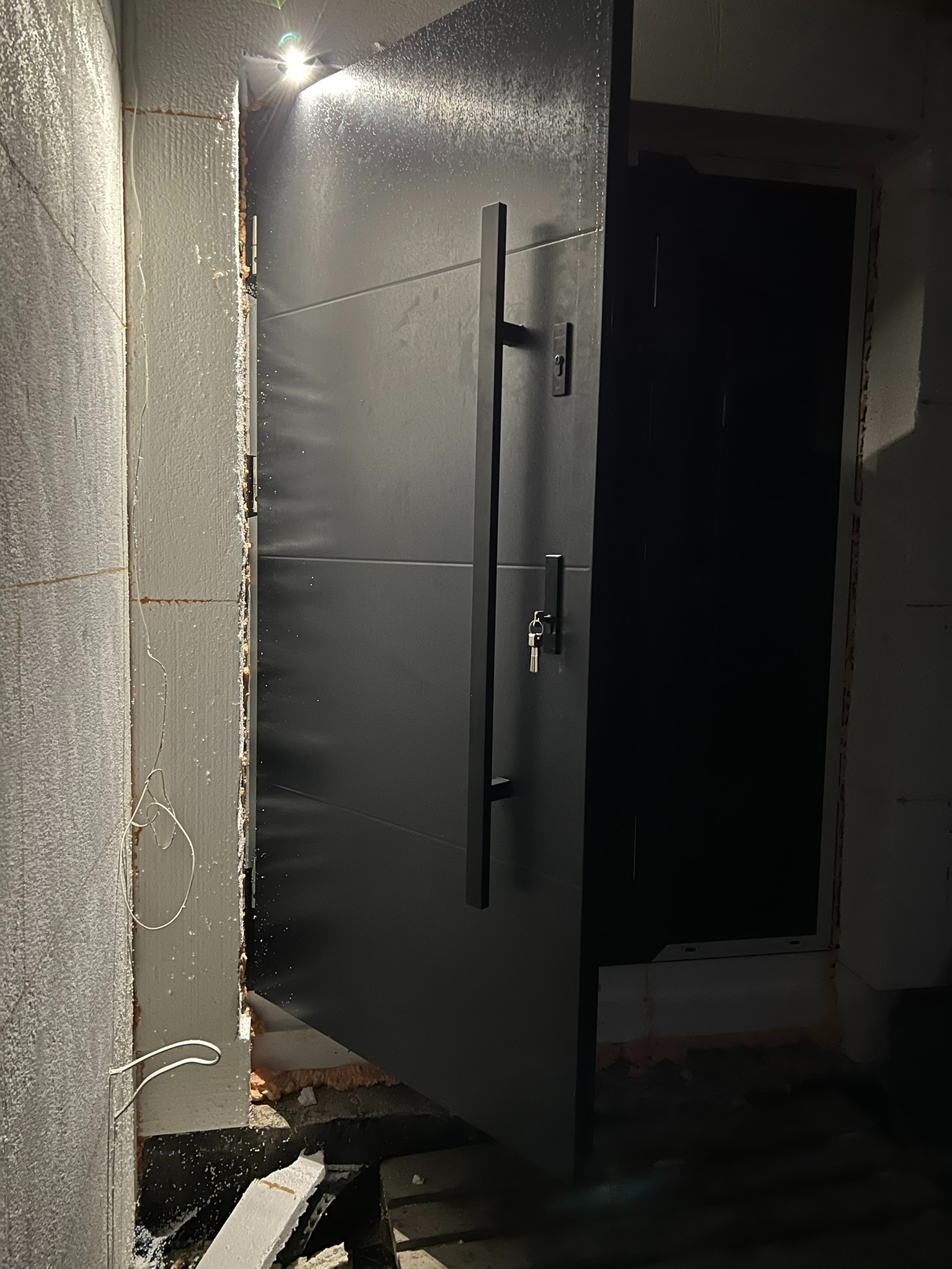 Ciemne drzwi zewnętrzne, częściowo otwarte, z widocznym zamkiem i kluczem, w trakcie montażu, z oświetleniem punktowym od góry i surowym wykończeniem ściany.