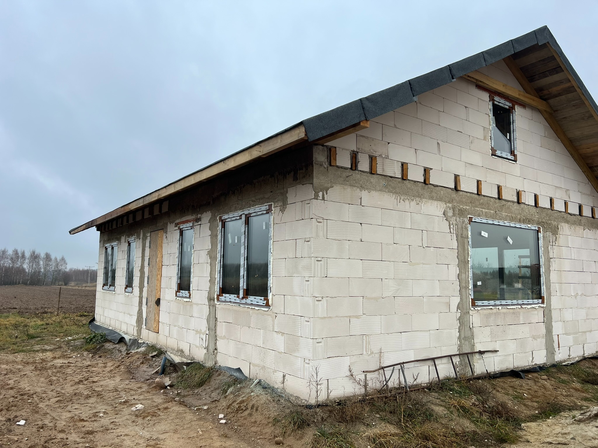Surowy stan domu z białego pustaka, z zamontowanymi oknami PCV i przygotowanym dachem. Widoczna drabina oparta o ścianę. Krajobraz polny w tle, pochmurne niebo.