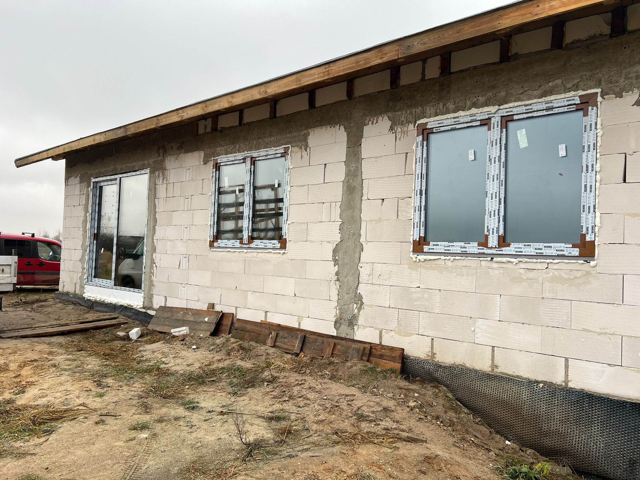 Ściana z betonu komórkowego z trzema oknami PCV w brązowych ramach, zabezpieczonymi folią. Widoczna warstwa izolacyjna przy gruncie. Budynek w trakcie budowy.