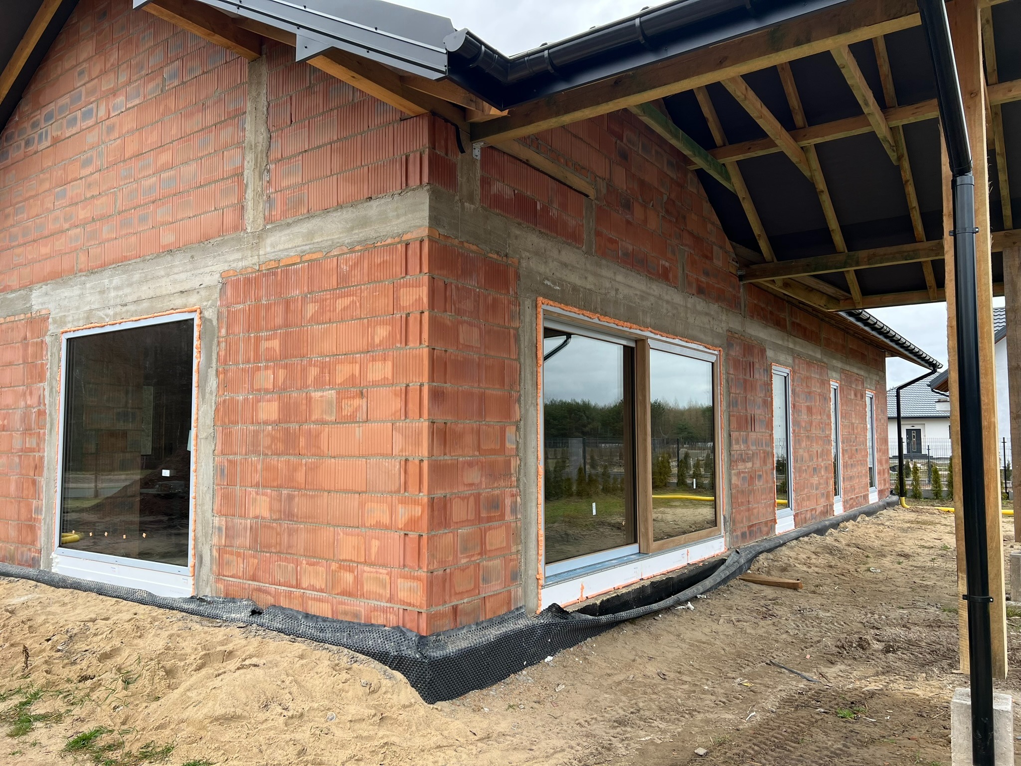Surowy budynek z czerwonej cegły z zamontowanymi oknami. Widoczna konstrukcja dachu i czarne rynny. Teren wokół budynku piaszczysty.