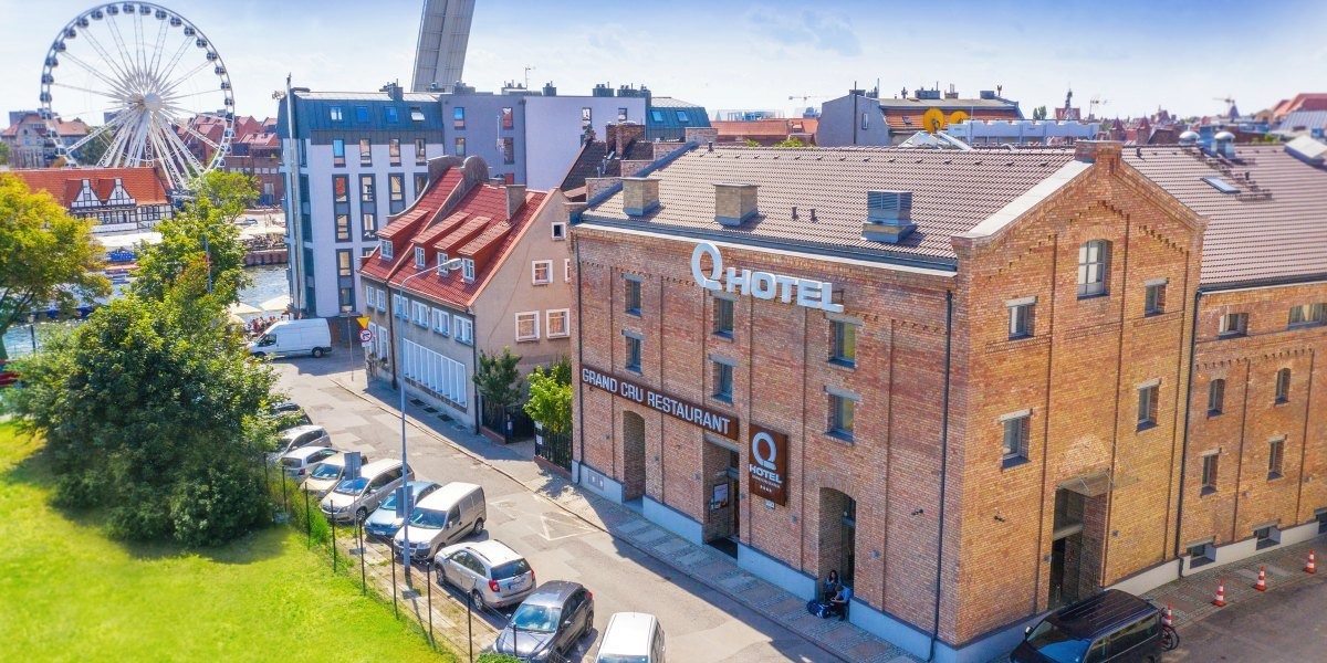 Widok z góry na ceglany budynek z napisem 'Q Hotel' i restauracją 'Grand Cru Restaurant', z zaparkowanymi samochodami wzdłuż ulicy i kołem młyńskim w tle.