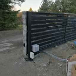 Nowoczesna, przesuwna brama wjazdowa z automatyką FAAC, zamontowana na posesji z widocznymi drzewami w tle, z elementami instalacji elektrycznej widocznymi obok napędu.