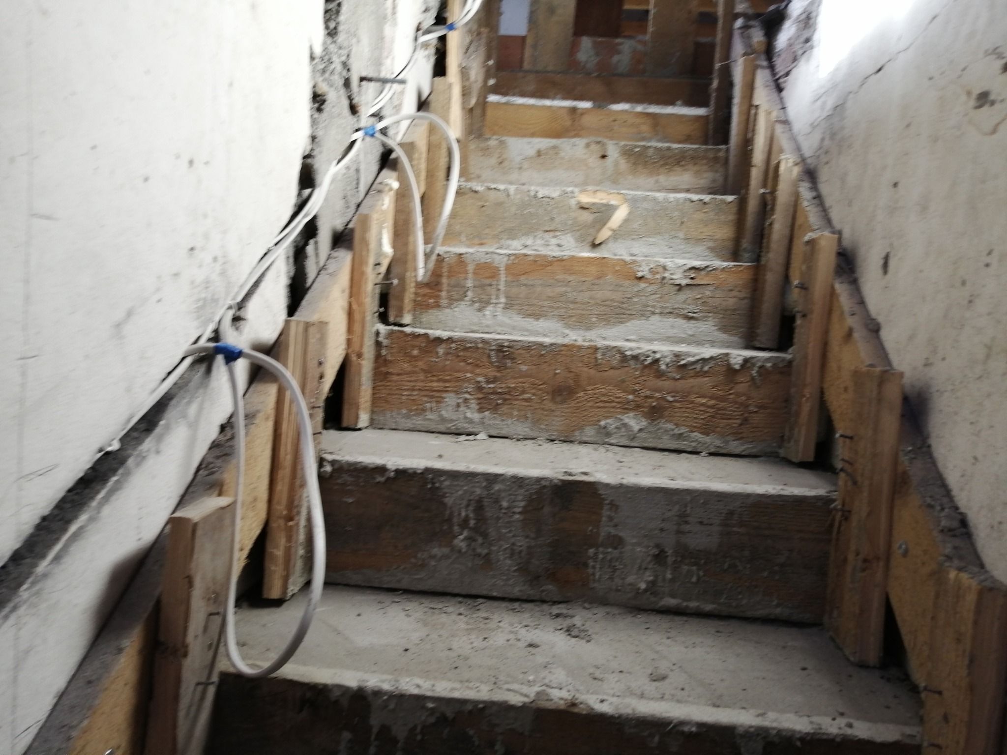 Kable elektryczne przymocowane do ściany wzdłuż surowych, drewnianych schodów w trakcie remontu. Widoczne ślady zaprawy i niedokończone wykończenie.