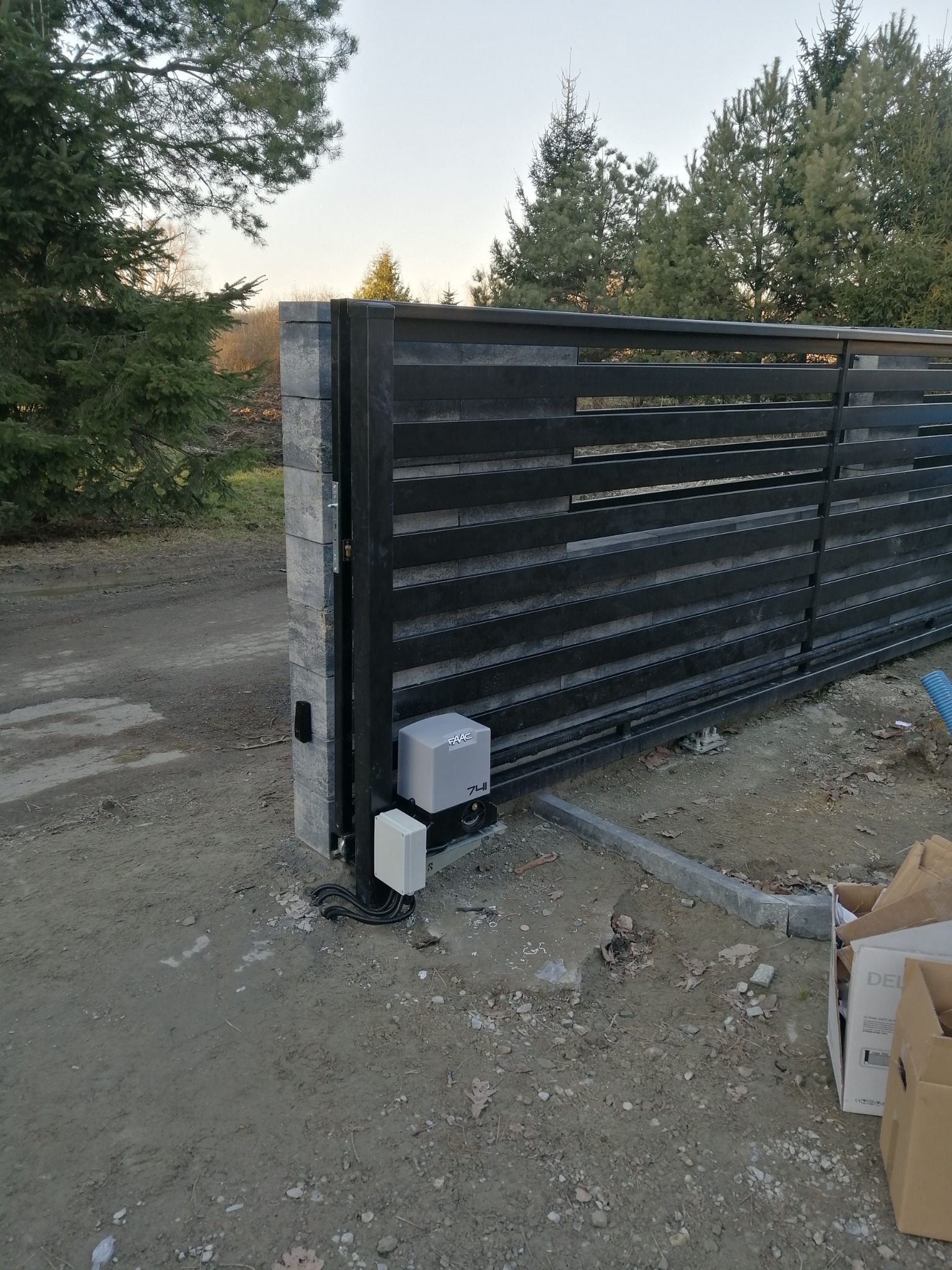 Nowoczesna, przesuwna brama wjazdowa z automatyką FAAC, zamontowana na posesji z widocznymi drzewami w tle, z elementami instalacji elektrycznej widocznymi obok napędu.