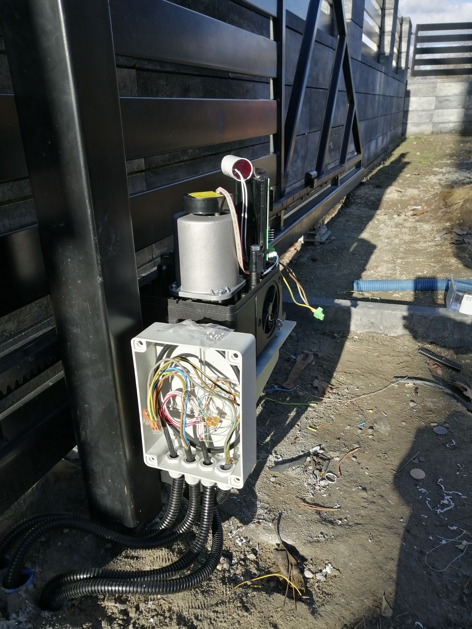 Szczegółowe ujęcie mechanizmu otwierającego przesuwną bramę wjazdową, z widoczną otwartą puszką elektryczną i podłączonymi przewodami.