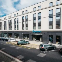 Elewacja nowoczesnego, kilkupiętrowego budynku z dużymi oknami, z niebieskim logo firmy nad wejściem i na ścianie, zaparkowane samochody wzdłuż ulicy, flaga powiewająca na wietrze.