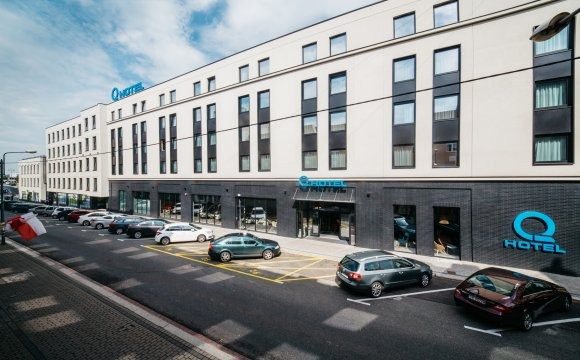 Elewacja nowoczesnego, kilkupiętrowego budynku z dużymi oknami, z niebieskim logo firmy nad wejściem i na ścianie, zaparkowane samochody wzdłuż ulicy, flaga powiewająca na wietrze.