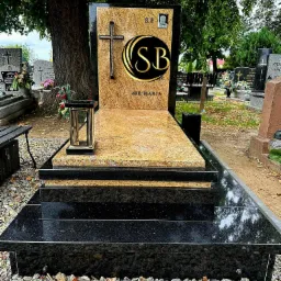 Nowoczesny nagrobek z czarnego i żółtego granitu, z krzyżem i inicjałami SB, otoczony kamieniami i innymi grobami na cmentarzu.