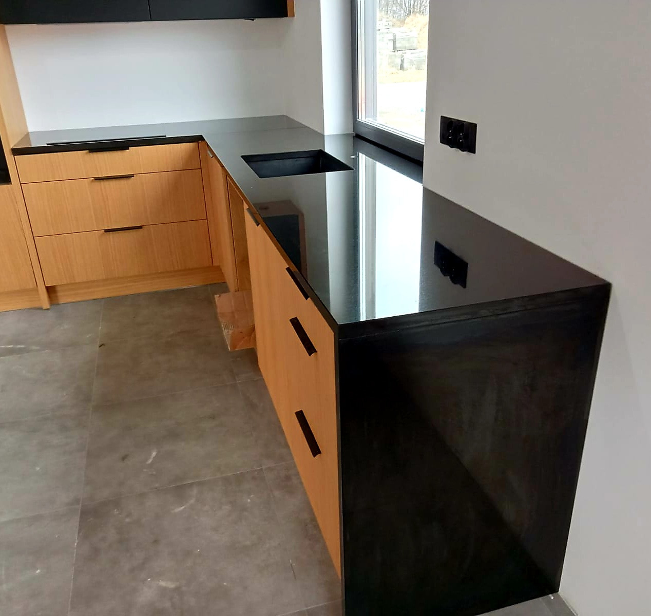 Czarny, błyszczący blat kamienny z wbudowanym zlewem, zamontowany w jasnej kuchni z drewnianymi szafkami i szarą podłogą. Widoczne gniazdka elektryczne na ścianie.