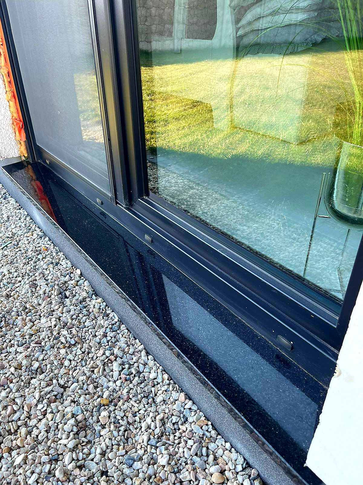 Czarny parapet zewnętrzny z konglomeratu, widok z bliska na drobny żwir u podstawy okna, odbicie trawnika w szybie.