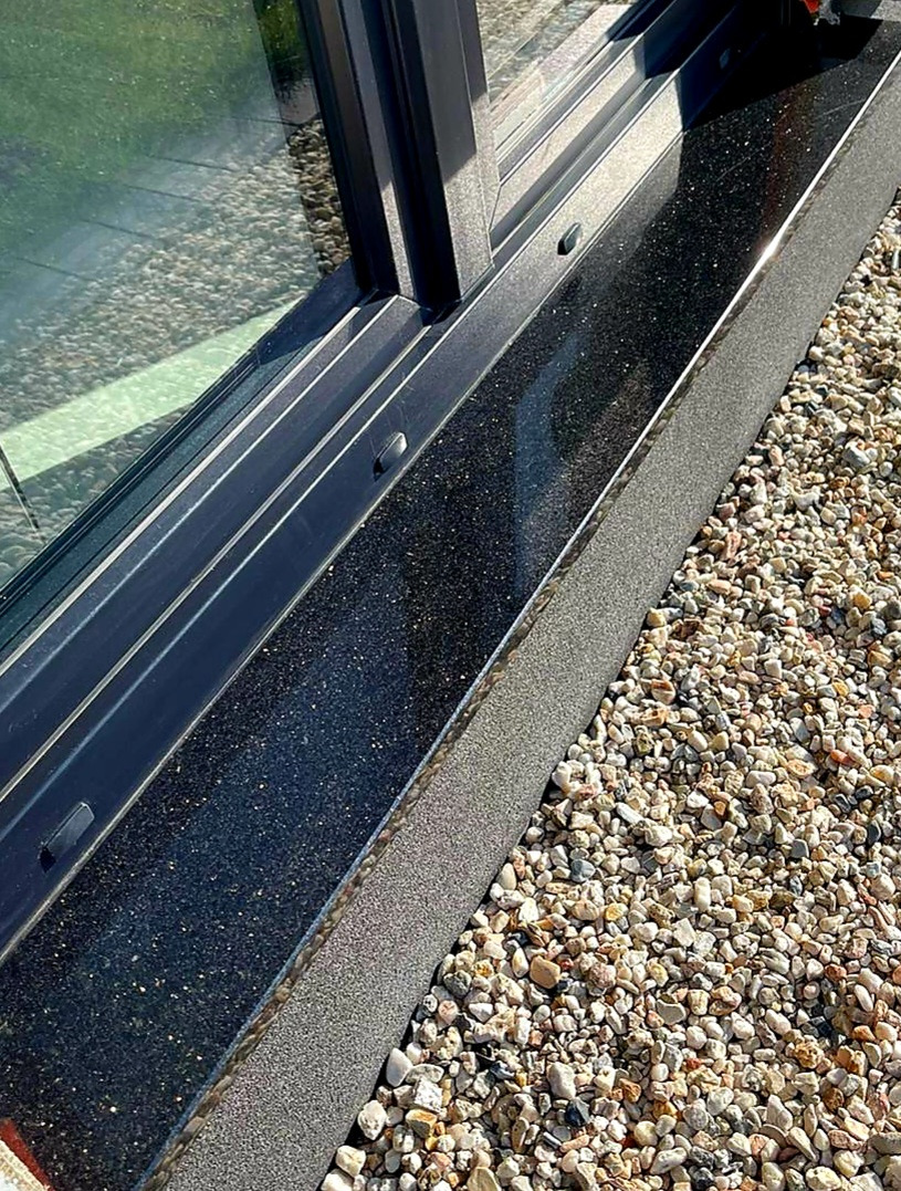 Zewnętrzny parapet z czarnego, błyszczącego kamienia z drobinkami, zamontowany pod oknem z ciemną ramą, otoczony drobnymi kamykami.