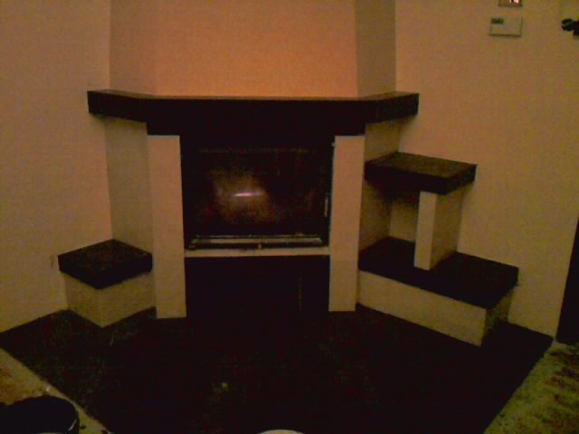 Kątowy kominek z ciemnym obramowaniem i półkami po bokach, wykończony w kolorach beżu i czerni, z widocznym paleniskiem i elementami instalacji.