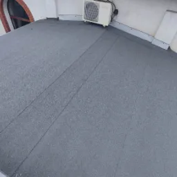 Płaski dach pokryty papą termozgrzewalną z widocznym klimatyzatorem na dachu, fragmentem elewacji z oknami w ceglanej oprawie i zielenią.