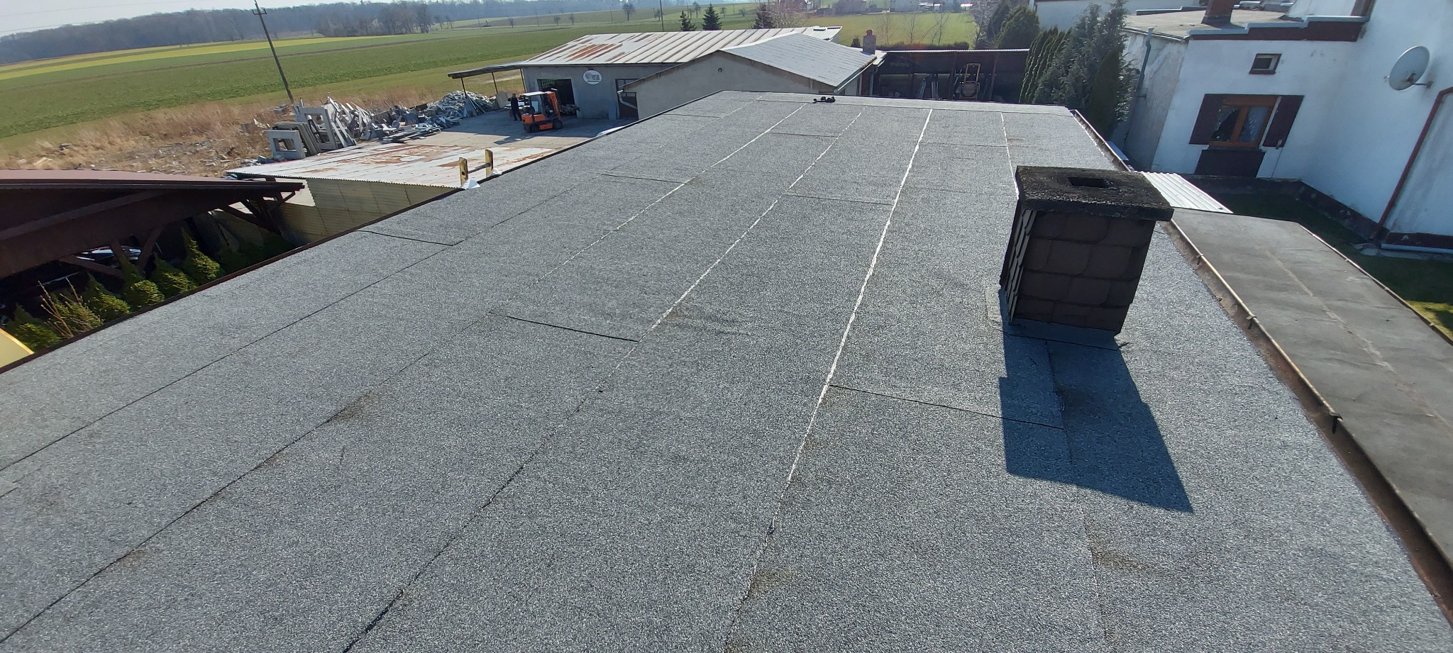 Widok z góry na płaski dach pokryty papą termozgrzewalną z widocznym kominem i elementami otoczenia.