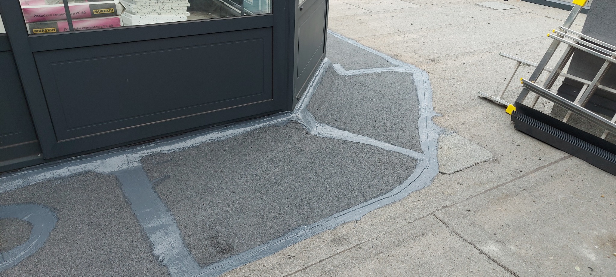 Uszczelnienie połączenia dachu z budynkiem przy użyciu szarej masy, z widoczną drabiną w tle.