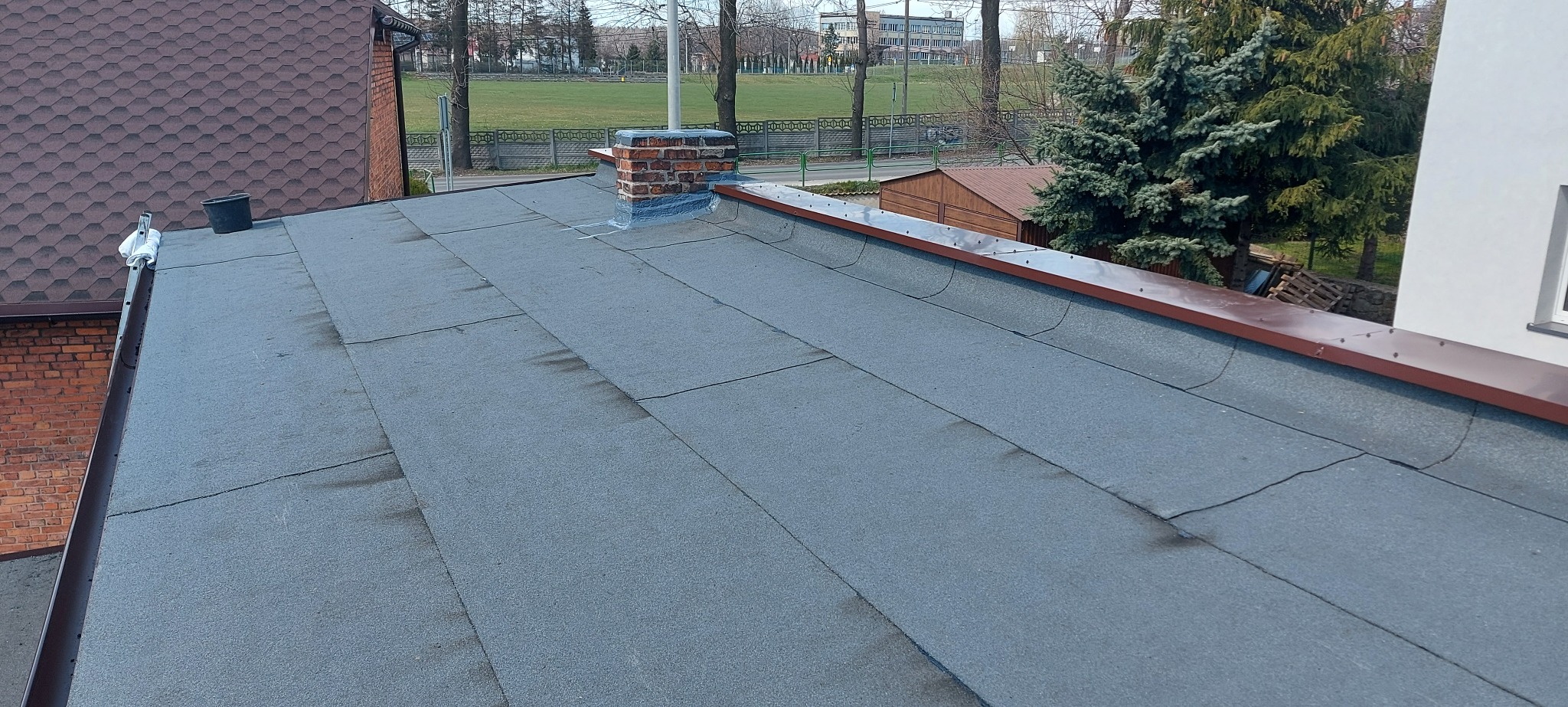 Szary dach pokryty papą z widocznym kominem obłożonym blachą, rynnami, obróbkami blacharskimi oraz elementami zgrzewania papy