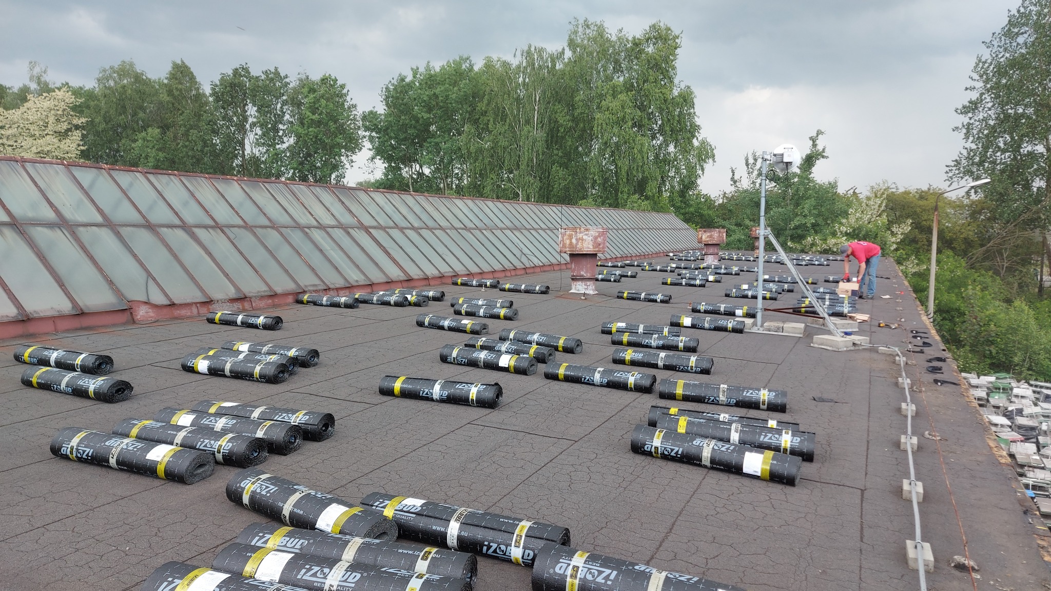 Rozwinięte rolki papy termozgrzewalnej marki IZOBud na płaskim dachu podczas prac dekarskich, w tle przeszklona konstrukcja dachu przemysłowego i pracujący dekarz.