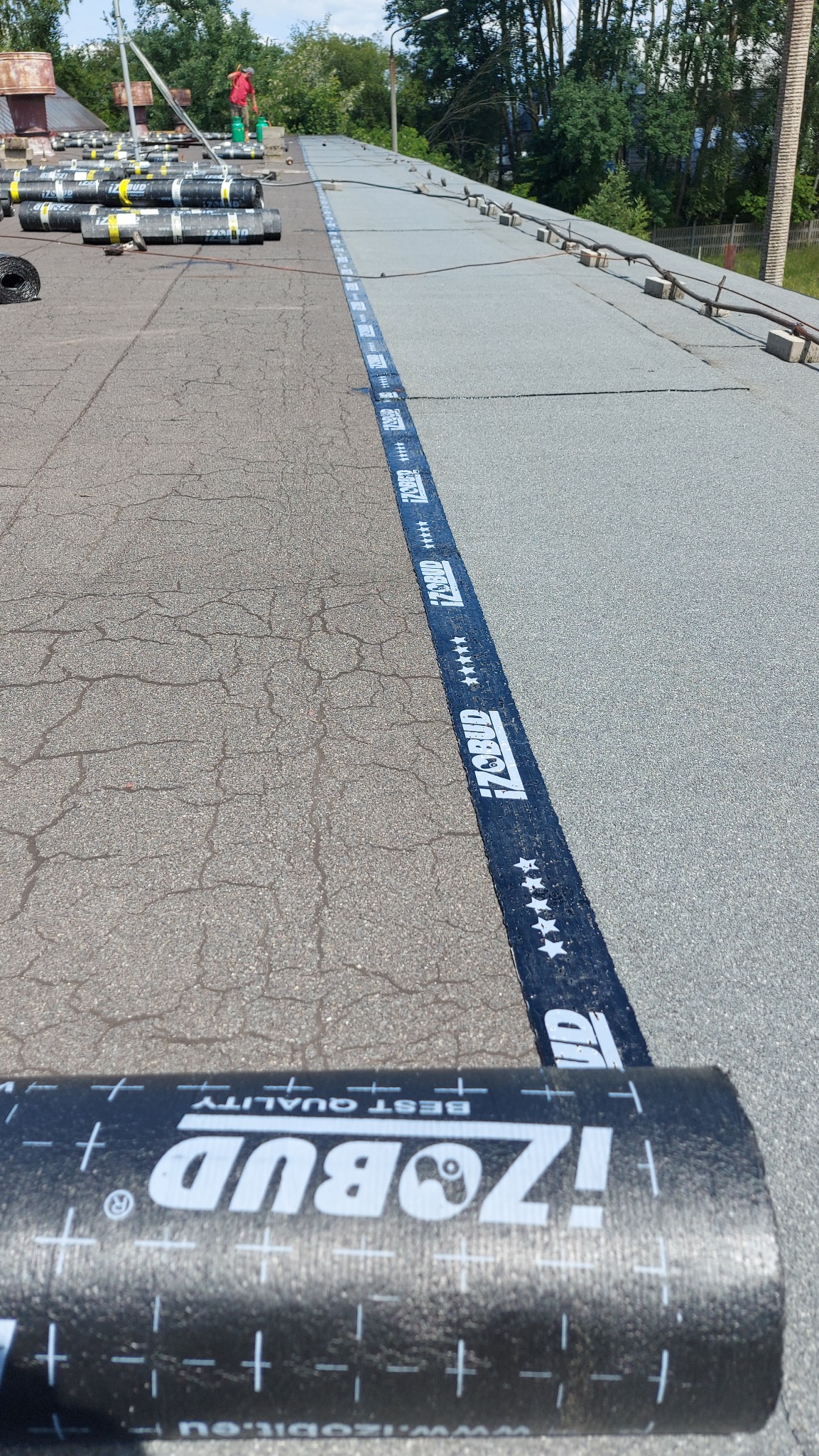 Dach pokryty papą termozgrzewalną z widocznym pasem z logo producenta, rolki papy czekają na montaż, pracownik w oddali.