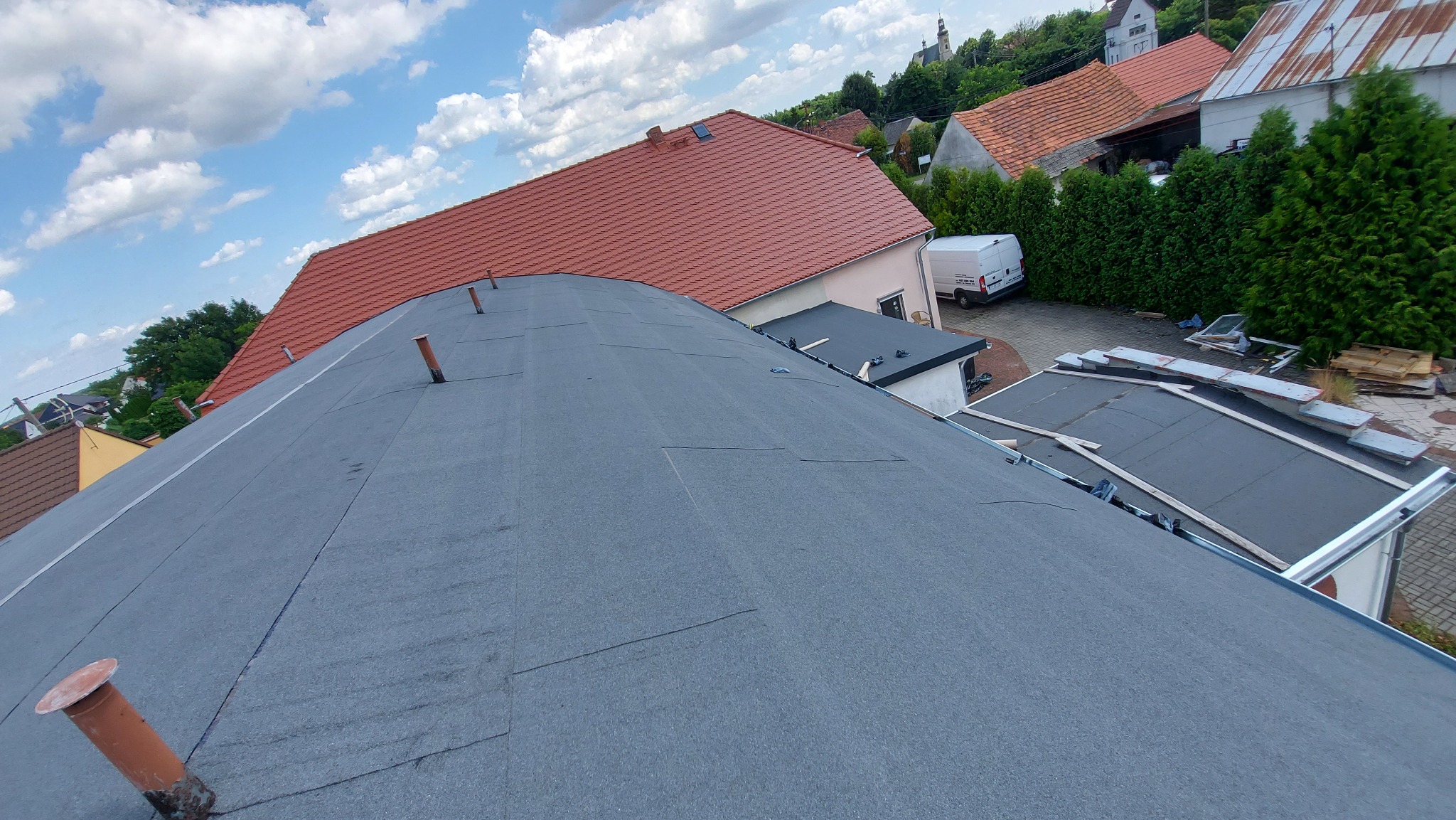Widok z góry na płaski dach pokryty papą termozgrzewalną, widoczne kominki wentylacyjne, w tle budynki mieszkalne i błękitne niebo z chmurami.