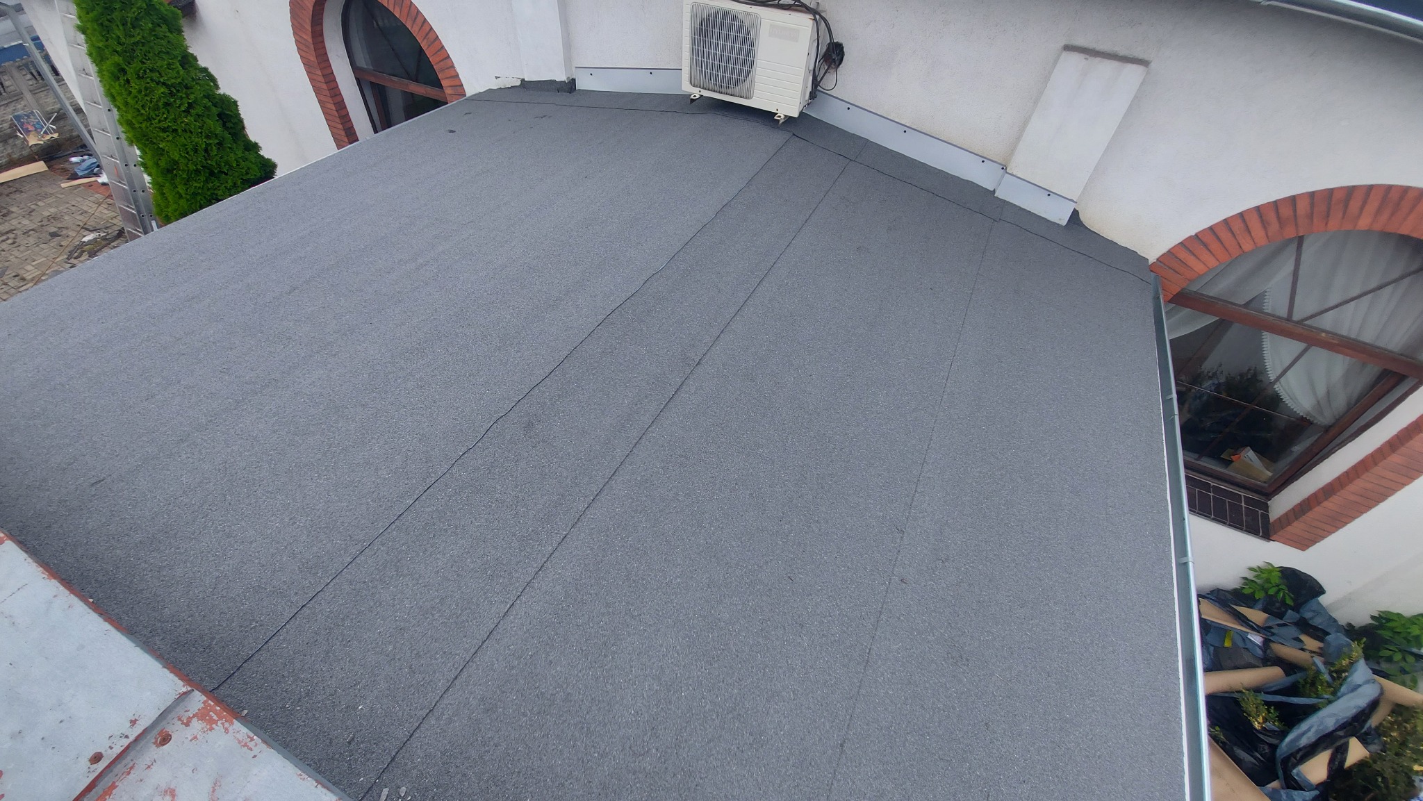 Płaski dach pokryty papą termozgrzewalną z widocznym klimatyzatorem na dachu, fragmentem elewacji z oknami w ceglanej oprawie i zielenią.