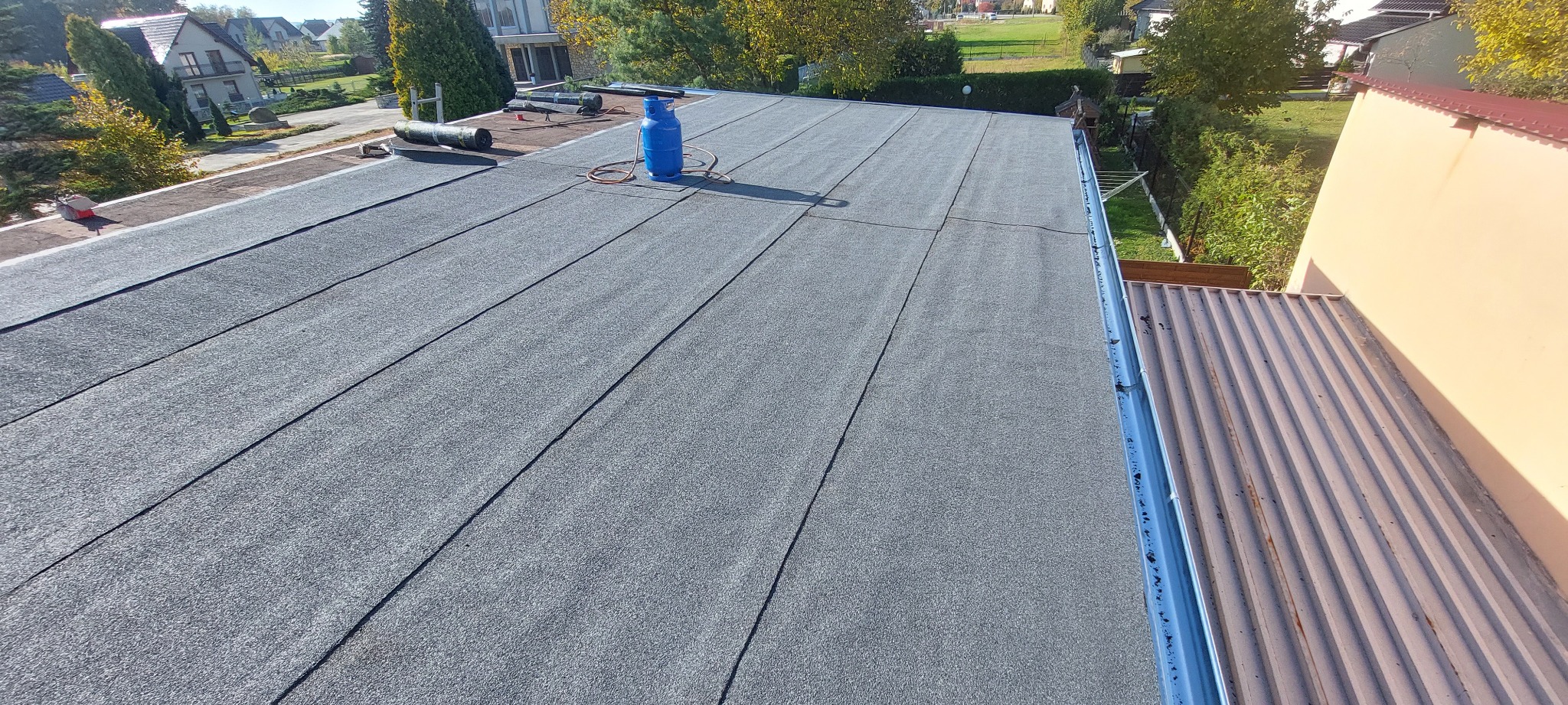 Widok z góry na płaski dach pokryty papą termozgrzewalną, z widoczną butlą gazową i rolkami papy oraz rynną w tle.