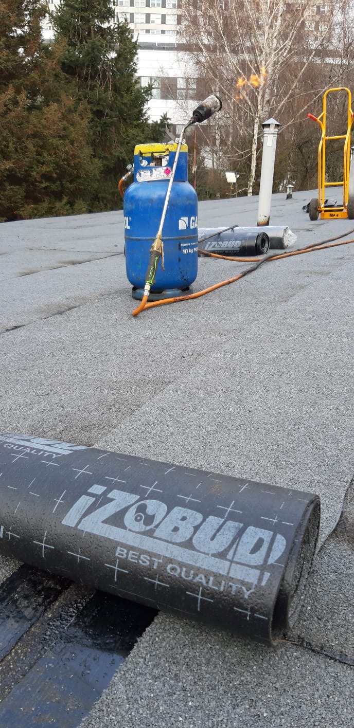 Zgrzewanie papy termozgrzewalnej marki IZObud na dachu z użyciem palnika gazowego i butli, widoczny wózek transportowy w tle.