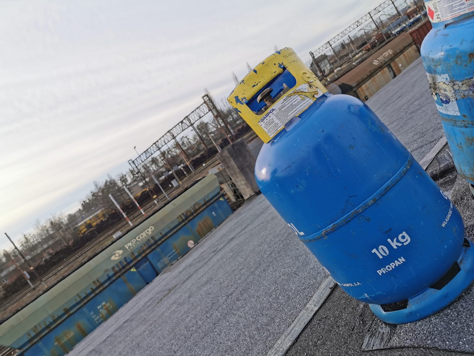 Dwie niebieskie butle z gazem propan-butan 10 kg na szarym dachu z widokiem na tory kolejowe i wagony PKP Cargo w tle.