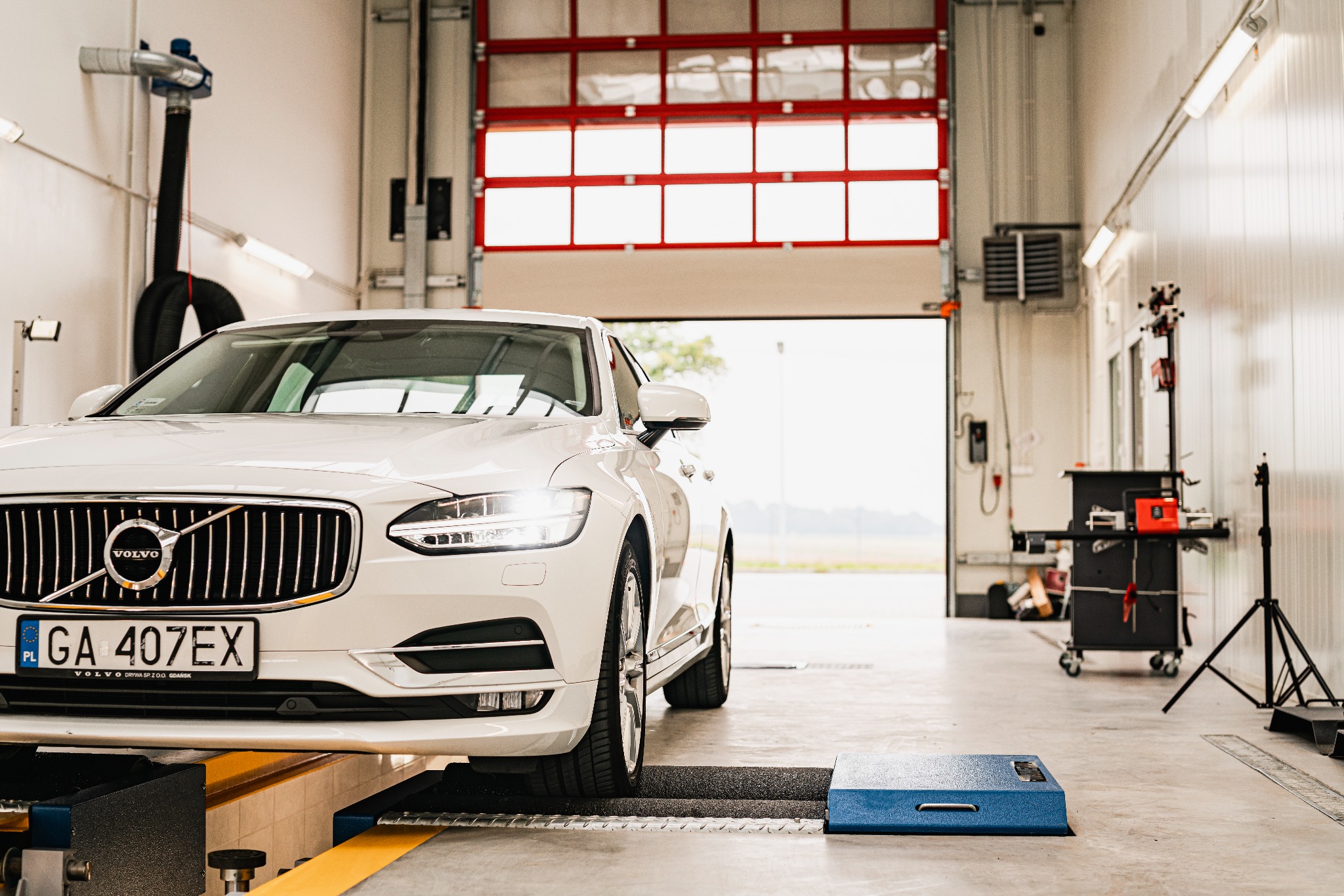 Biały samochód Volvo na podnośniku w warsztacie, widoczne otwarte wrota garażowe i sprzęt diagnostyczny w tle.