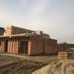 Budowa domu murowanego z czerwonej cegły, widoczne ściany w trakcie wznoszenia, drewniane podpory i rusztowania, hałda piasku na pierwszym planie.